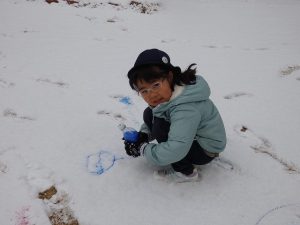 雪の上に女の子がペットボトルに入った絵の具で絵を書いてる